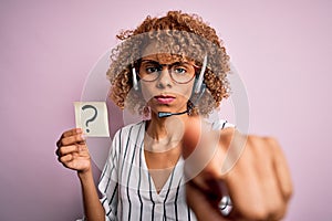 African american call center agent woman using headset holding paper with question mark pointing with finger to the camera and to