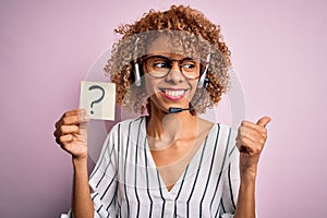 African american call center agent woman using headset holding paper with question mark pointing and showing with thumb up to the
