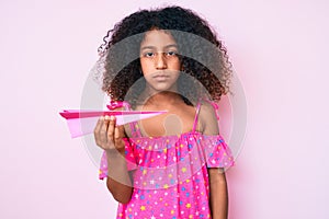 African american child with curly hair holding paper airplane thinking attitude and sober expression looking self confident