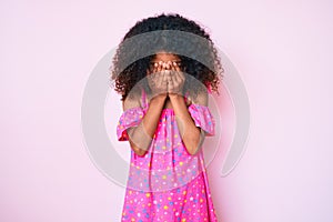 African american child with curly hair wearing casual dress with sad expression covering face with hands while crying