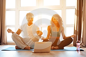 African American couple doing yoga exercises at home. They sit on the floor on yoga mats in a lotus position.