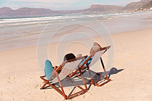 African american couple relaxing on deckchairs at sandy beach during sunny day, copy space