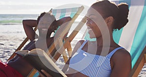 African american couple sunbathing and reading book while sitting on deck chairs at the beach