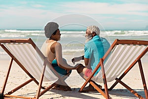 African american couple talking while sitting on deckchairs at sandy beach against cloudy sky