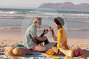 African american couple toasting juice while sitting with food on blanket against sea at sunset