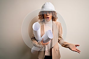 African american curly architect woman wearing safety helmet and glasses holding blueprints scared in shock with a surprise face,