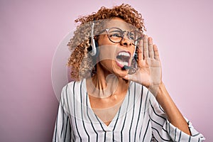 African american curly call center agent woman working using headset over pink background shouting and screaming loud to side with