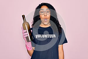 African american curly woman wearing volunteer t-shirt doing volunteering recycling glass bottle scared in shock with a surprise