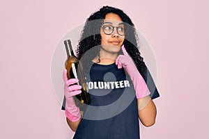 African american curly woman wearing volunteer t-shirt doing volunteering recycling glass bottle serious face thinking about