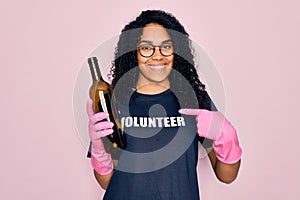 African american curly woman wearing volunteer t-shirt doing volunteering recycling glass bottle with surprise face pointing
