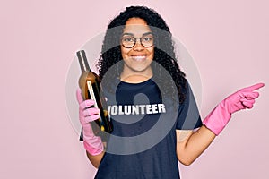 African american curly woman wearing volunteer t-shirt doing volunteering recycling glass bottle very happy pointing with hand and