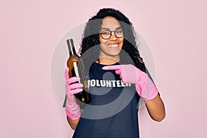 African american curly woman wearing volunteer t-shirt doing volunteering recycling glass bottle very happy pointing with hand and