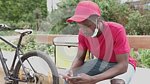 African american delivery man using mobile while sitting on public park