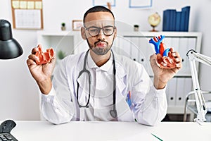 African american doctor man holding anatomical heart at the clinic puffing cheeks with funny face