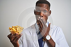 African american doctor man holding bowl with macaroni pasta over isolated white background cover mouth with hand shocked with