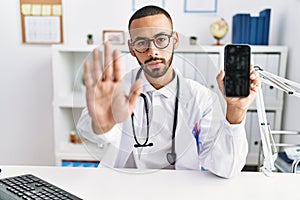 African american doctor man holding smartphone showing blank screen at the clinic with open hand doing stop sign with serious and