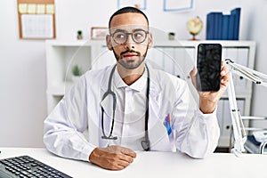 African american doctor man holding smartphone showing blank screen at the clinic thinking attitude and sober expression looking