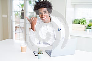 African American doctor man working with laptop at the clinic pointing and showing with thumb up to the side with happy face