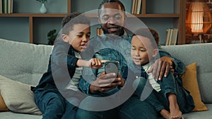 African American ethnic father man dad teach little boys brothers siblings children using mobile phone sitting on sofa