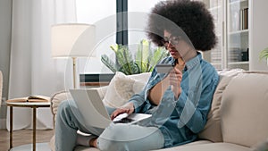 African American ethnic girl making online payment holding bank credit card using modern laptop at home sitting on sofa