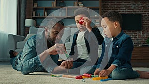 African American ethnic man father with two little preschool sons boys children play game lying on floor at home dad