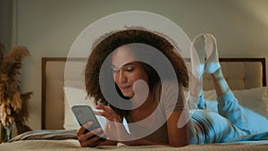African American ethnic woman 20s girl curly hair scrolling mobile phone relaxing at home evening night insomnia smiling
