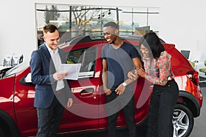 African american family at car dealership. Salesman is showing trunk of new red car