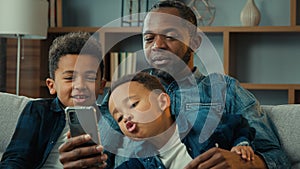 African American family father man and little kids sons boys children sit on sofa look at mobile screen having fun using