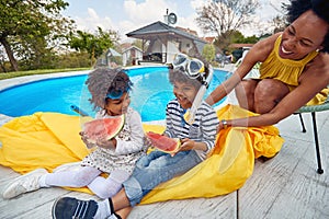 African american family outdoors by pool, mother with her two children, a boy and a girl eating watermelon fruit having fun