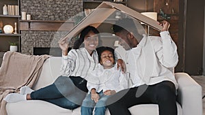 African american family parents with daughter little girl adopted child sit on sofa under cardboard roof dreaming about