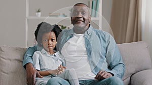 African american family sitting on sofa kid girl waving hello greeting adult father hugging daughter talking at camera