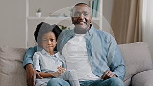 African american family sitting on sofa kid girl waving hello greeting adult father hugging daughter talking at camera