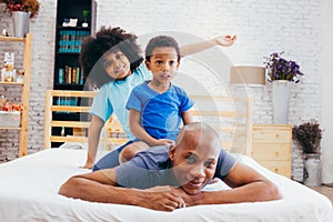 African American family of three, kids sitting on father`s back at home