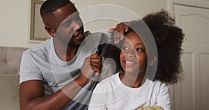 African american father brushing his daughters hair while sitting on the bed at home