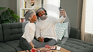 African american father and daughter wearing king crown and baggy eyes pad make selfie by smartphone at home