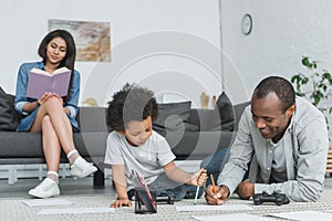 african american father drawing with son and mother reading book on sofa