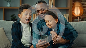 African American father man with little boys children sitting together on sofa couch using smartphone mobile phone