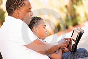 African American Father and Mixed Race Son Using Computer Tablet on Bench in Park