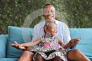 African American father playing with his little daughter who sitting on his lap while spending happy time together at home on sofa