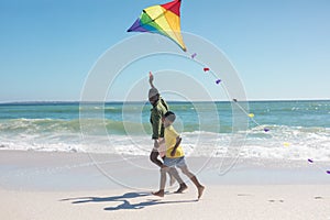 African american father and son holding hands while running with kite at beach on sunny day