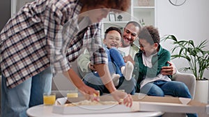 African american father tickling two sons on couch smiling mother cutting pizza