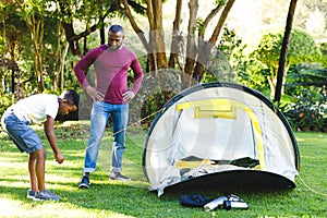 African american father watching son pitching tent in sunny garden