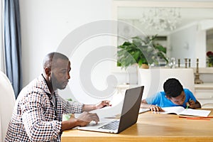 African american father working on laptop in dining room with son sitting with him doing homework