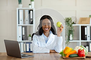 African American female dietitian holding apple and smiling at camera in modern office