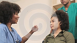 African American female doctor checks patient girl in wheelchair at hospital.