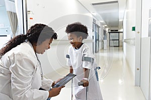 African american female doctor and girl patient with crutches using tablet in corridor at hospital