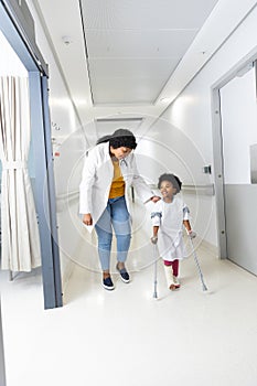 African american female doctor and girl patient walking with crutches in corridor at hospital
