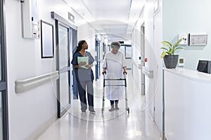 African american female doctor walking with senior female patient with crutches in hospital corridor