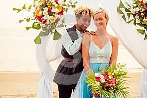 African american female groom in black suit and caucasian bride in dress in ceremony on tropical beach under wedding