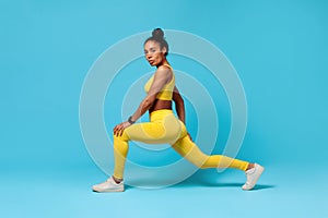 African American fitness lady stretching legs lunging forward, blue background